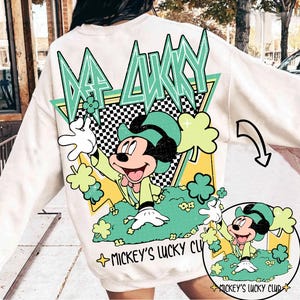 Pode incluir: Camisola creme com um desenho do Mickey Mouse vestido de duende, rodeado por trevos. O texto "BE LUCKY" está acima do desenho, com "Mickey's Lucky Club" abaixo. O design inclui detalhes em verde, amarelo e preto.
