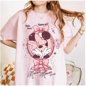 May include: A pale pink t-shirt with a Minnie Mouse design. The graphic shows Minnie in a mirror, bows, and the text "Hello, there! Happy girl! Minnie Mouse xoxo." Additional text reads "Why settle for less fun?"