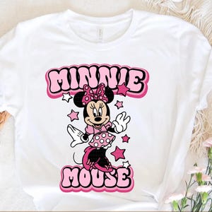 Peut inclure: T-shirt blanc avec un dessin de Minnie Mouse en robe et n&oelig;ud rose. Le texte "MINNIE MOUSE" est affich&eacute; au-dessus et en dessous du personnage dans un style de lettres bulle avec des accents roses et blancs. Des &eacute;toiles roses entourent le personnage.