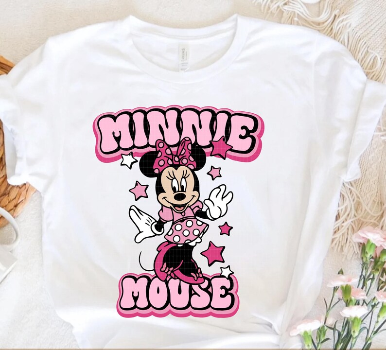 Peut inclure: T-shirt blanc avec un dessin de Minnie Mouse en robe et n&oelig;ud rose. Le texte "MINNIE MOUSE" est affich&eacute; au-dessus et en dessous du personnage dans un style de lettres bulle avec des accents roses et blancs. Des &eacute;toiles roses entourent le personnage.