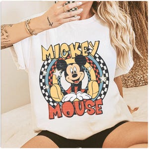 May include: White t-shirt with a retro Mickey Mouse design. The graphic features Mickey's image, the words "Mickey Mouse" in a colourful, vintage font, and a chequered pattern. The shirt is a classic, casual style.