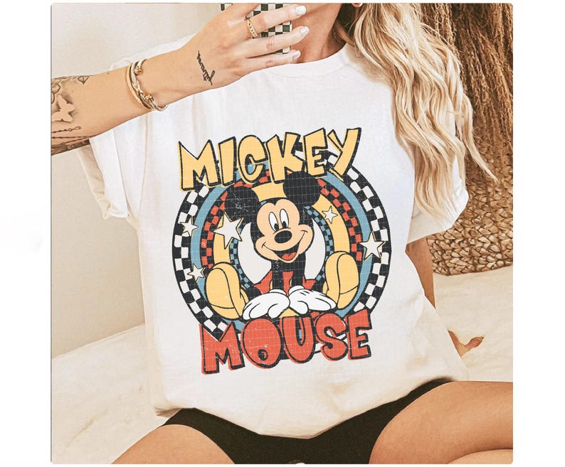 May include: White t-shirt with a retro Mickey Mouse design. The graphic features Mickey's image, the words "Mickey Mouse" in a colourful, vintage font, and a chequered pattern. The shirt is a classic, casual style.