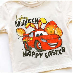 May include: Cream-colored t-shirt featuring a red cartoon car with bunny ears, resembling Lightning McQueen, and the text "Happy Easter." The car has the number "95" on it, and is surrounded by Easter eggs.