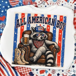 Op de afbeelding: Wit T-shirt met een afbeelding van een wasbeer met zonnebril, bandana en hoed met de tekst "USA". De wasbeer zit in een bruine fauteuil, houdt een biertje vast en heeft cowboylaarzen voor zich. De tekst "ALL AMERICAN BOY" staat erboven.
