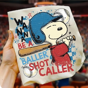 May include: Off-white t-shirt featuring a cartoon Snoopy character in a baseball helmet and uniform, holding a bat. The shirt has the text "Wanna Be A Baller Shot Caller" in blue and red. Baseball-themed apparel.