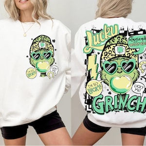 K&ouml;nnte beinhalten: Wei&szlig;es Sweatshirt mit einem Grinch-&auml;hnlichen Charakter, der eine herzf&ouml;rmige Sonnenbrille, einen Leopardenhut und eine Kaugummiblase tr&auml;gt. Der Text enth&auml;lt "I'm so lucky" und "Lucky Me!". Auf der R&uuml;ckseite steht "Lucky" und "Grinch".