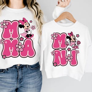 May include: White sweatshirts with pink text and Minnie Mouse graphics. One sweatshirt says "MAMA" and the other says "MINI". The text is in a bubble font with a black outline, and the Minnie Mouse graphic is in pink and black.