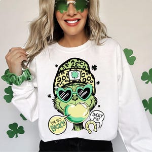 K&ouml;nnte beinhalten: Wei&szlig;es Sweatshirt mit einer gr&uuml;nen Cartoonfigur mit herzf&ouml;rmiger Sonnenbrille und Leopardenmuster-Hut. Die Figur hat Sprechblasen mit der Aufschrift "I'M SO LUCKY!" und "LUCKY ME!". Kleeblattdekorationen sind im Hintergrund.