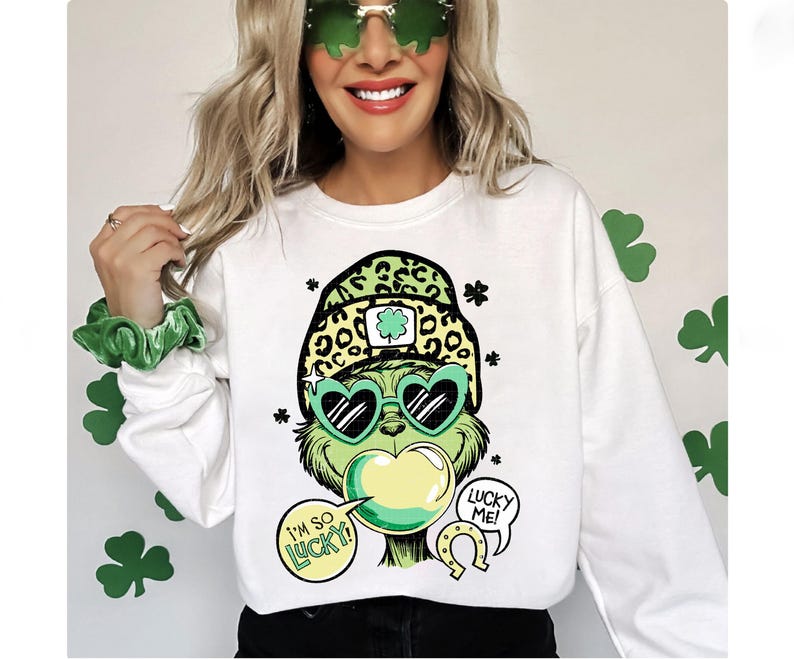 K&ouml;nnte beinhalten: Wei&szlig;es Sweatshirt mit einer gr&uuml;nen Cartoonfigur mit herzf&ouml;rmiger Sonnenbrille und Leopardenmuster-Hut. Die Figur hat Sprechblasen mit der Aufschrift "I'M SO LUCKY!" und "LUCKY ME!". Kleeblattdekorationen sind im Hintergrund.