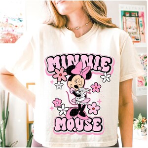 May include: Cream-colored t-shirt featuring a graphic of Minnie Mouse holding a small gray rabbit, surrounded by pink and white flowers. The words "MINNIE MOUSE" are in a bubble-style font, outlined in black.