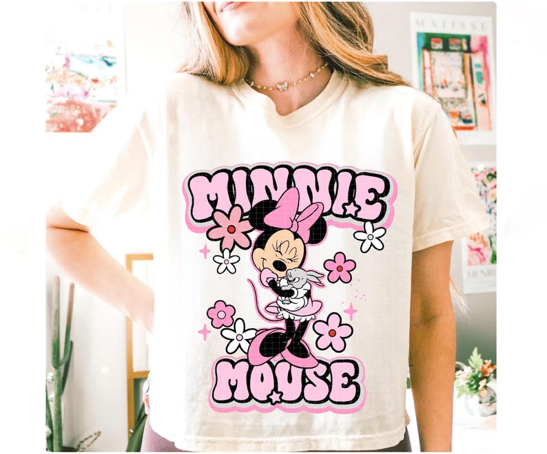 May include: Cream-colored t-shirt featuring a graphic of Minnie Mouse holding a small gray rabbit, surrounded by pink and white flowers. The words "MINNIE MOUSE" are in a bubble-style font, outlined in black.