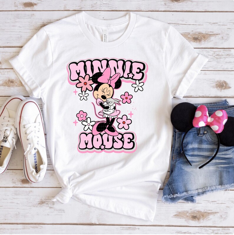 May include: White t-shirt featuring a Minnie Mouse graphic in pink and black. The design includes the words "MINNIE MOUSE" in a retro font, surrounded by pink flowers. The shirt is paired with white sneakers and denim shorts.