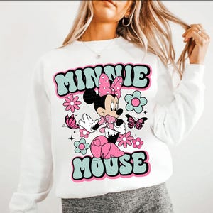 May include: White sweatshirt featuring a retro design of Minnie Mouse. The design includes the words "MINNIE MOUSE" in a teal and black font, along with pink flowers, butterflies, and Minnie Mouse in a pink polka dot dress.