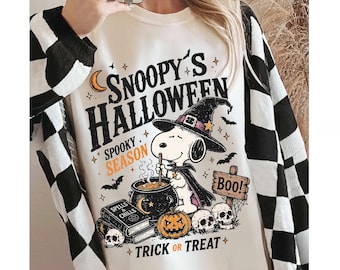 May include: A cream-colored sweatshirt featuring a graphic print of Snoopy wearing a witch hat and cape, stirring a cauldron. The text reads SNOOPY'S HALLOWEEN, SPOOKY SEASON, BOO, and TRICK OR TREAT, accompanied by illustrations of skulls, a pumpkin, and books.