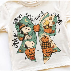Puede incluir: Una camiseta color crema con un gráfico de un lazo verde azulado que muestra a Snoopy y Woodstock. El diseño incluye calabazas, hojas de otoño y el texto "It's the most boo-tiful time of the year" en letra negra. Estética de estilo vintage.