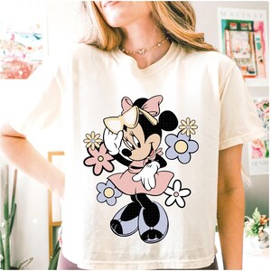 May include: Cream-colored t-shirt featuring a Minnie Mouse graphic. Minnie is depicted in a pink dress, bow, and sunglasses, surrounded by pastel flowers. The design is a playful and colorful illustration.