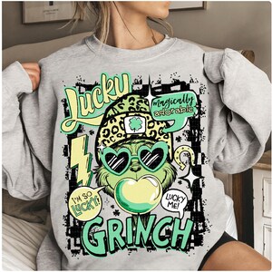 K&ouml;nnte beinhalten: Ein hellgraues Sweatshirt mit einem gr&uuml;nen Grinch-Charakter, der eine herzf&ouml;rmige Sonnenbrille und einen Leopardenhut tr&auml;gt. Das Design enth&auml;lt die W&ouml;rter "Lucky Grinch" und "I'm so lucky!" in einer verspielten Schriftart. Das Gesamtthema ist St. Patrick's Day.