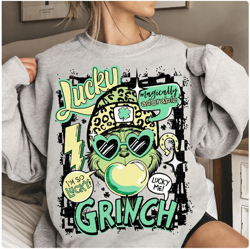 K&ouml;nnte beinhalten: Ein hellgraues Sweatshirt mit einem gr&uuml;nen Grinch-Charakter, der eine herzf&ouml;rmige Sonnenbrille und einen Leopardenhut tr&auml;gt. Das Design enth&auml;lt die W&ouml;rter "Lucky Grinch" und "I'm so lucky!" in einer verspielten Schriftart. Das Gesamtthema ist St. Patrick's Day.