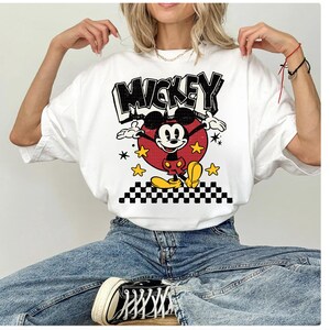 Puede incluir: Camiseta blanca con un gr&aacute;fico de Mickey Mouse y la palabra "Mickey" en negro. Mickey est&aacute; en un c&iacute;rculo rojo con estrellas amarillas y un borde a cuadros blanco y negro.