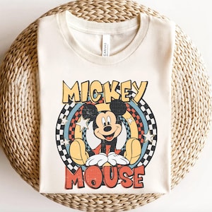 May include: Cream-coloured t-shirt featuring a retro design of Mickey Mouse. The graphic includes the words "MICKEY MOUSE" in a vintage font, with Mickey sitting inside a circular design with stars and a checkered pattern.