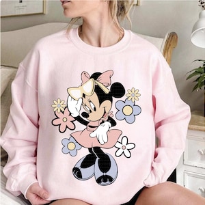 May include: A light pink sweatshirt featuring a Minnie Mouse graphic. The cartoon character is surrounded by pastel-colored flowers. Minnie Mouse is wearing a pink dress, white gloves, and black shoes. A large bow adorns her head.