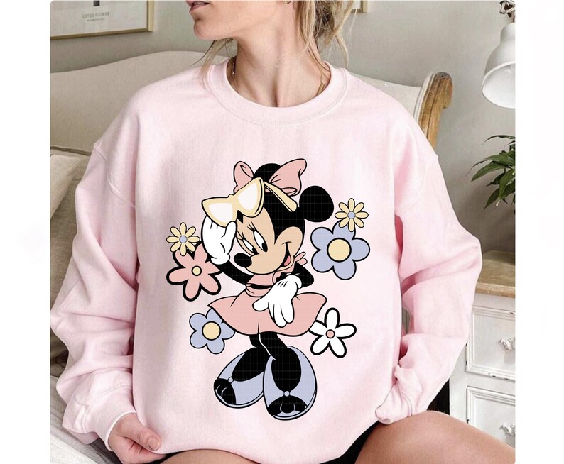 May include: A light pink sweatshirt featuring a Minnie Mouse graphic. The cartoon character is surrounded by pastel-colored flowers. Minnie Mouse is wearing a pink dress, white gloves, and black shoes. A large bow adorns her head.