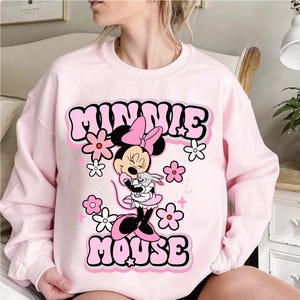 May include: A light pink sweatshirt featuring a graphic of Minnie Mouse holding a small gray bunny. The words "MINNIE MOUSE" are in a bubble font with a pink outline. Pink and white flowers and stars surround the graphic.