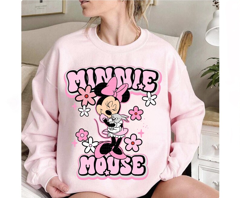 May include: A light pink sweatshirt featuring a graphic of Minnie Mouse holding a small gray bunny. The words "MINNIE MOUSE" are in a bubble font with a pink outline. Pink and white flowers and stars surround the graphic.