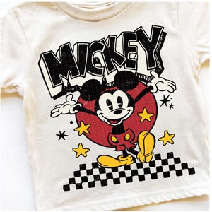 Puede incluir: Camiseta color crema con un dise&ntilde;o de Mickey Mouse de estilo vintage. El dise&ntilde;o incluye la palabra "MICKEY" en letras negras, Mickey Mouse en un c&iacute;rculo rojo, estrellas amarillas y un patr&oacute;n de cuadros blanco y negro.