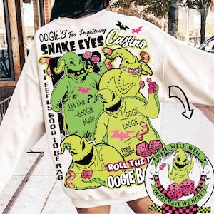 May include: Cream-colored sweatshirt featuring a graphic of the character Oogie Boogie from The Nightmare Before Christmas. The design includes text that reads "Oogie's Snake Eyes Casino" and "Roll the Oogie Boogie". The graphic is in shades of green, pink, and black.
