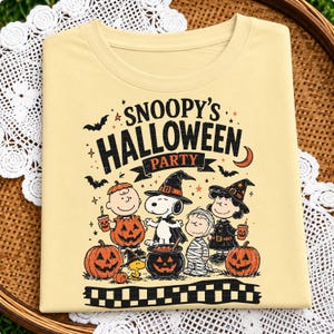 May include: A pale yellow t-shirt featuring a graphic print with the text SNOOPY'S HALLOWEEN PARTY. The design includes Snoopy, Charlie Brown, and other characters in costumes, surrounded by pumpkins, bats, stars, and a crescent moon.