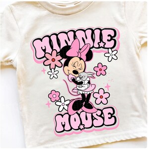 May include: Cream-colored t-shirt featuring a graphic of Minnie Mouse in a pink dress and bow, hugging a small gray bunny. The words "MINNIE MOUSE" are in pink bubble letters, surrounded by pink and white flowers.