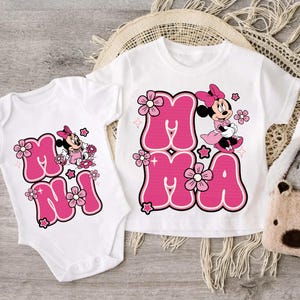 Puede incluir: Body y camiseta blancos con un diseño rosa de "Minnie". El diseño incluye la palabra "MAMA" en letras rosas con burbujas, Minnie Mouse y detalles florales. La camiseta y el body se muestran sobre una superficie de madera clara.