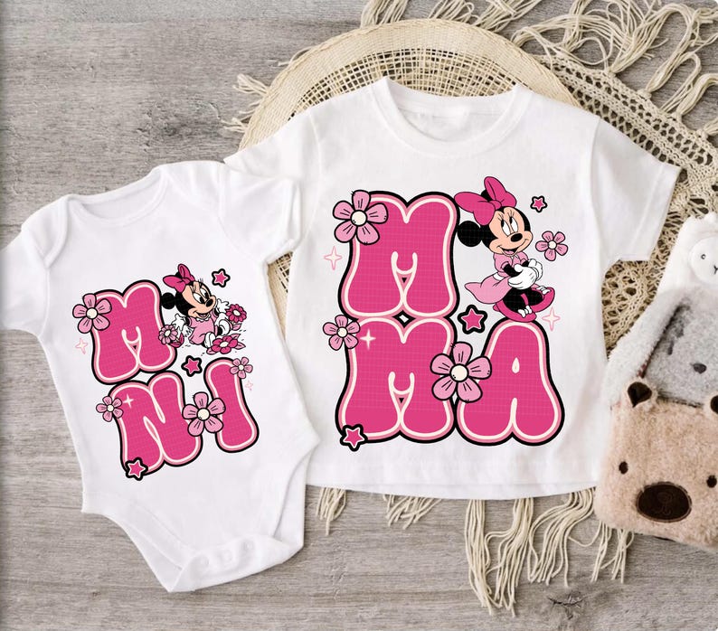May include: White baby onesie and t-shirt with a pink "Minnie" graphic. The graphic includes the word "MAMA" in pink bubble letters, Minnie Mouse, and floral accents. The t-shirt and onesie are displayed on a light wooden surface.
