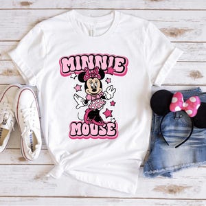 Peut inclure: T-shirt blanc avec un motif de Minnie Mouse rose et le texte "MINNIE MOUSE". L'image comprend des baskets blanches, un short en jean et des oreilles de Minnie avec un n&oelig;ud rose.