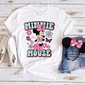May include: White t-shirt featuring a retro-style graphic of Minnie Mouse with the text "MINNIE MOUSE" in a bubble font. The design includes pink and blue flowers and butterflies. The image also shows white sneakers, denim shorts, and a Minnie Mouse ear headband.