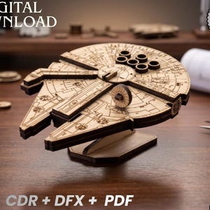Könnte beinhalten: Ein Holzmodell des Raumschiffs Millennium Falcon, als digitaler Laser-Cut-Datei-Download. Das hellbraune Modell weist detaillierte kreisförmige und rechteckige Elemente auf. Der Text "DIGITALER DOWNLOAD" und "LASER CUT DATEI" sind im Bild enthalten.