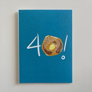Könnte beinhalten: Eine Grußkarte mit türkisblauem Hintergrund zeigt die Zahl "4" und eine Pfannkuchen-Illustration mit einem Butterstück. Das Ausrufezeichen und die Zahl sind weiß. Der Pfannkuchen ist braun und gelb.
