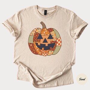 Könnte beinhalten: Ein sandfarbenes Kurzarm-T-Shirt mit einer großen zentralen Grafik eines Kürbisses. Der Kürbis ist in einem Patchwork-Muster gestaltet, das aus verschiedenen herbstlichen Stoffen in den Farben Orange, Braun, Grün und Creme besteht.