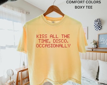 May include: A pale yellow short-sleeve boxy t-shirt hanging on a wooden hanger. The shirt features red pixelated text on the front that reads: KISS ALL THE TIME. DISCO, OCCASIONALLY. The garment has a crew neckline and a relaxed, loose-fitting cut.
