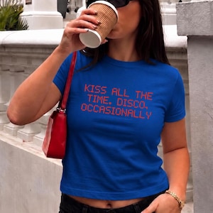 May include: A royal blue short-sleeved t-shirt featuring the orange pixelated text "KISS ALL THE TIME. DISCO. OCCASIONALLY" printed on the chest. The shirt has a crew neckline and a cropped fit, paired with black denim pants and a red shoulder bag.