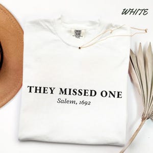 May include: A white short-sleeved t-shirt with a crew neckline and a Comfort Colors brand label. The front features black text that reads "THEY MISSED ONE" in a bold serif font, followed by "Salem, 1692" in a smaller italicised serif font.