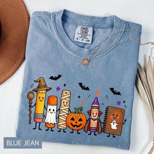 May include: A blue crew neck t-shirt featuring a graphic of anthropomorphic school supplies dressed in Halloween costumes, including a pencil witch, a glue stick ghost, a mummy ruler, a pumpkin, a vampire crayon, and a haunted notebook with bats.