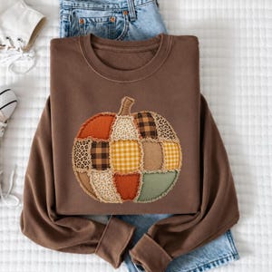 May include: A brown crewneck sweatshirt featuring a large, central pumpkin graphic composed of various fabric patches. The pumpkin design includes orange, yellow, brown, and green sections with plaid, leopard print, and floral patterns with frayed edges.