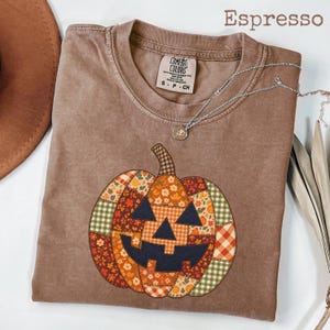 Könnte beinhalten: Ein espressofarbenes T-Shirt mit einer großen, zentralen Kürbisgrafik, die aus verschiedenen Patchwork-Mustern wie Blumen-, Karomustern und Vichy-Karos in Orange, Grün und Braun besteht. Das T-Shirt hat einen Rundhalsausschnitt und ein Comfort Colors-Etikett.