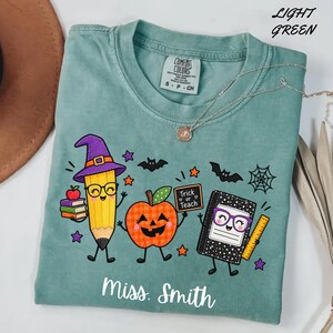 Op de afbeelding: Een lichtgroen t-shirt met een afbeelding van een potlood met een heksenhoed, een pompoenappel en een notitieboek met een bril. Het ontwerp bevat vleermuizen, een spinnenweb en een bordje met Trick or Teach. De tekst Miss Smith staat eronder.