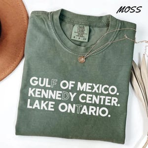 Op de afbeelding: Een mosgroen t-shirt met korte mouwen en witte tekst die luidt GULF OF MEXICO. KENNEDY CENTER. LAKE ONTARIO. Het shirt heeft een ronde hals en een Comfort Colors merklabel in de kraag. Een dunne goudkleurige ketting ligt op de stof.