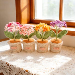 以下が含まれることがあります： ベージュの鉢に入った、かぎ針編みのフラワーアレンジメント4つ。花はピンク、オレンジ、紫の色合いで、緑の葉が付いています。鉢はスカラップエッジと小さなラベルが付いています。白いレースのテーブルクロスの上にアレンジされています。