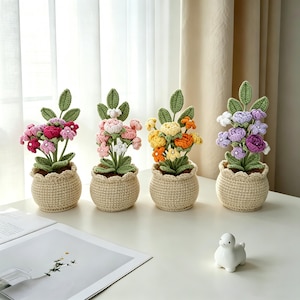 May include: Four crocheted potted flower arrangements featuring green leaves and stems in beige textured pots. The flowers are in shades of pink, light pink, yellow, orange, and purple, with small white accents. A small white ceramic duck figurine sits nearby.