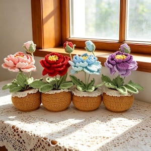 May include: Four crocheted flowers in round, tan, textured pots. Each pot features a white lace trim at the top. The flowers are pink, red, light blue, and purple, each with a yellow center, green leaves, and a small matching bud on a stem.